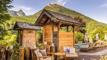 A wooden cabin with a pool area, surrounded by lush greenery and mountains.