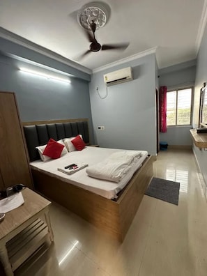 A bedroom with a bed, bedside table, wardrobe, ceiling fan, air conditioning unit, and a window with curtains. - Fabhotel Adhikari (Kolkata)