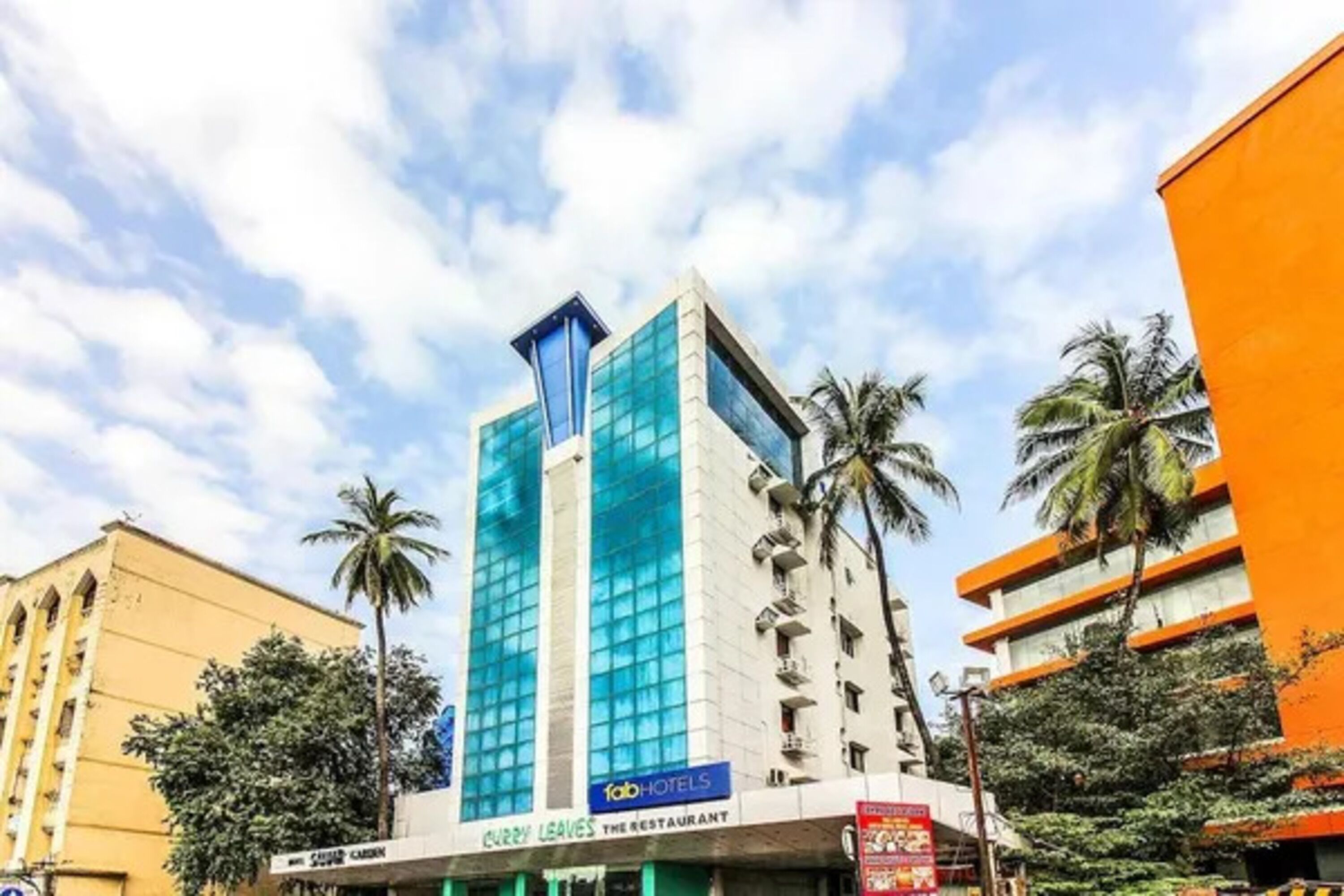 Un hotel alto con fachada de vidrio moderna, un cartel para el Restaurante Curry Leaves y palmeras al fondo.