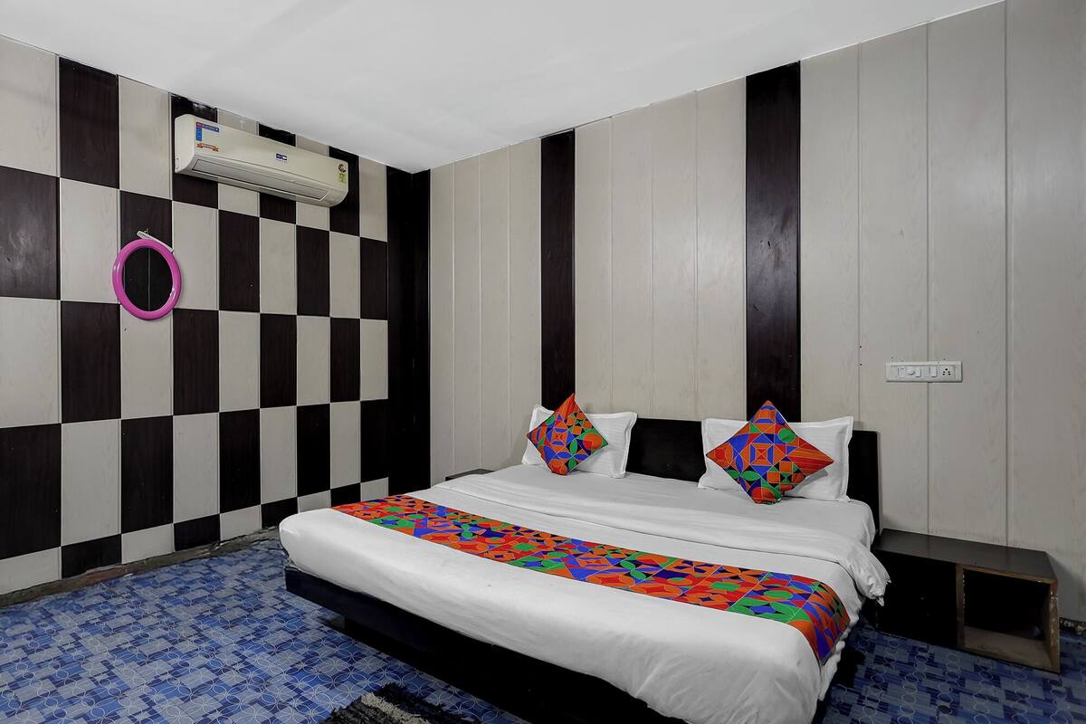 Een hotelkamer met een geruit patroon op de muur, een bed met witte beddengoed en kleurrijke kussens, een blauw patroon tapijt, en een airconditioner aan de muur.
