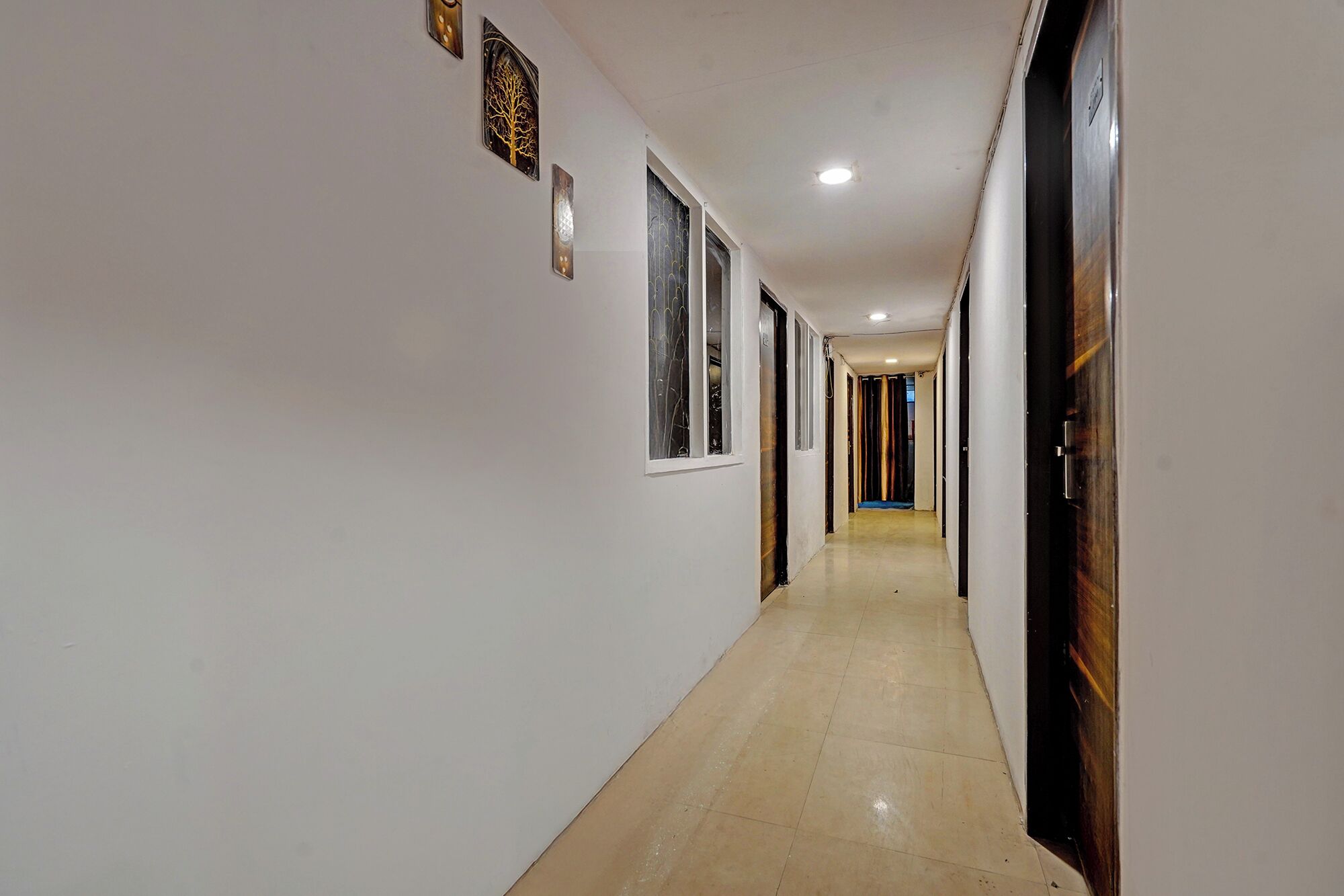 A long, narrow hallway with white walls and a tiled floor. There are several doors on the right side, and a few windows on the left.