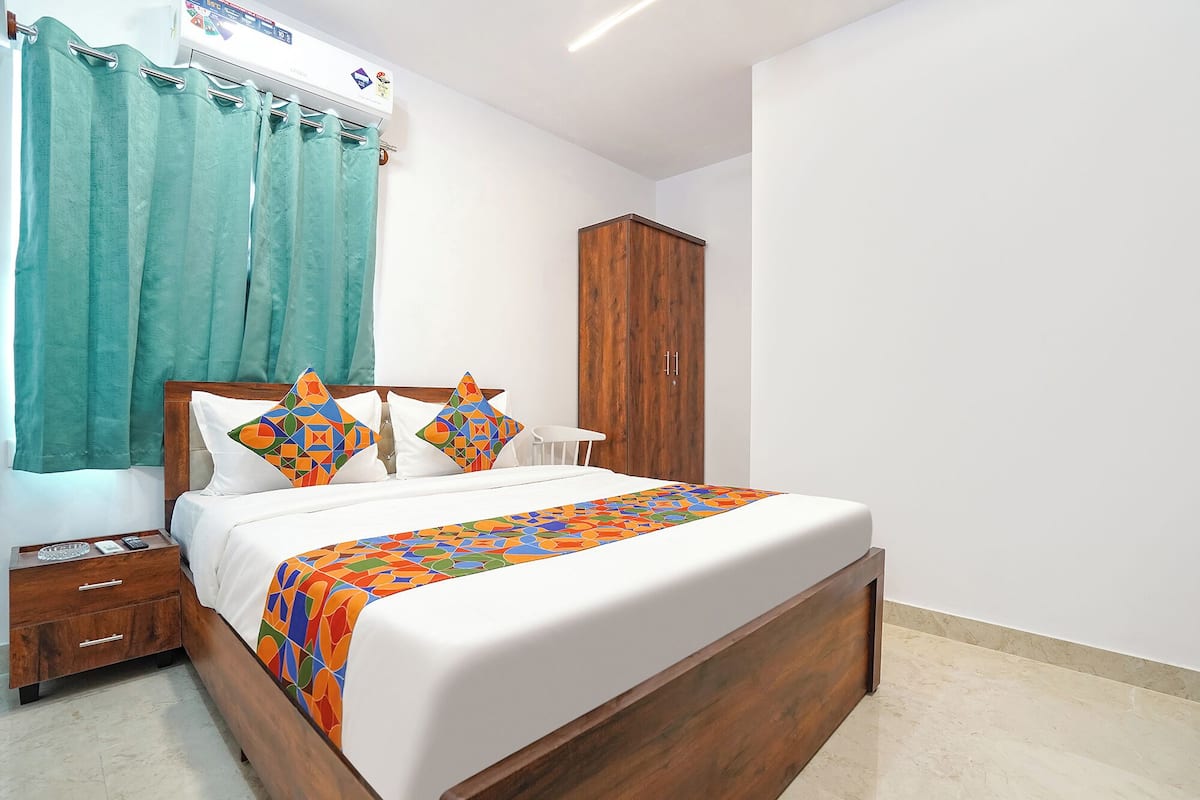 A neatly made bed with a colorful geometric-patterned bedspread, a wooden nightstand with a glass object, a white chair, and a wooden wardrobe.