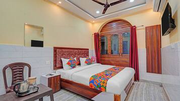 A hotel room with a bed, a wooden headboard, a nightstand, a table with a kettle, and a TV mounted on the wall.