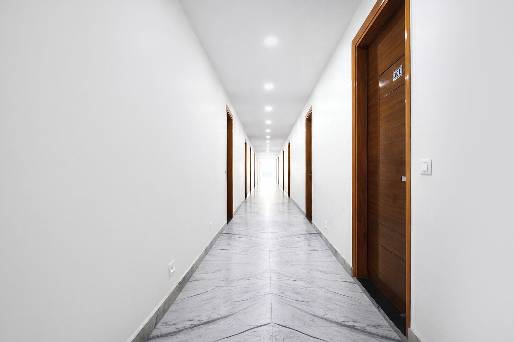 A long, narrow hallway with white walls and a marble floor, leading to a wooden door with a sign above it.