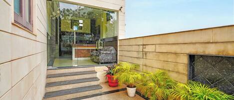 A modern building with a glass entrance, a small outdoor seating area with a sofa and a table, and several potted plants.