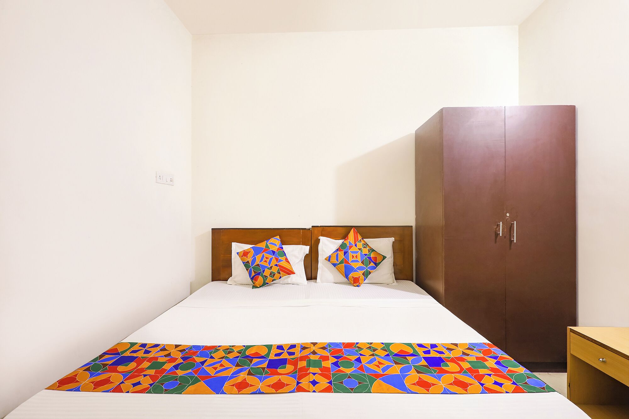 Ein Einzelbett mit einer farbenfrohen Bettdecke mit geometrischem Muster und zwei Kissen im ähnlichen Design. Eine Holzgarderobe befindet sich rechts neben dem Bett.