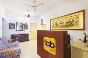 A hotel reception area with a wooden reception desk, a sofa, a dresser, and a ceiling fan. - Fabhotel Disha Inn (Chennai)