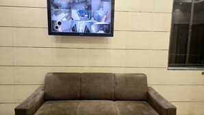 A brown leather sofa against a tiled wall with a flat-screen TV displaying security footage. - Fabhotel Kamal (Mumbai)