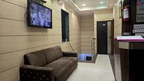 A lobby with a brown sofa, a television mounted on the wall, a reception desk, and a fire extinguisher. - Fabhotel Kamal (Mumbai)