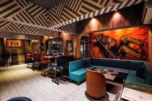 A restaurant with a geometric patterned ceiling, a mural of a guitarist, and a mix of seating including a blue sofa and chairs.