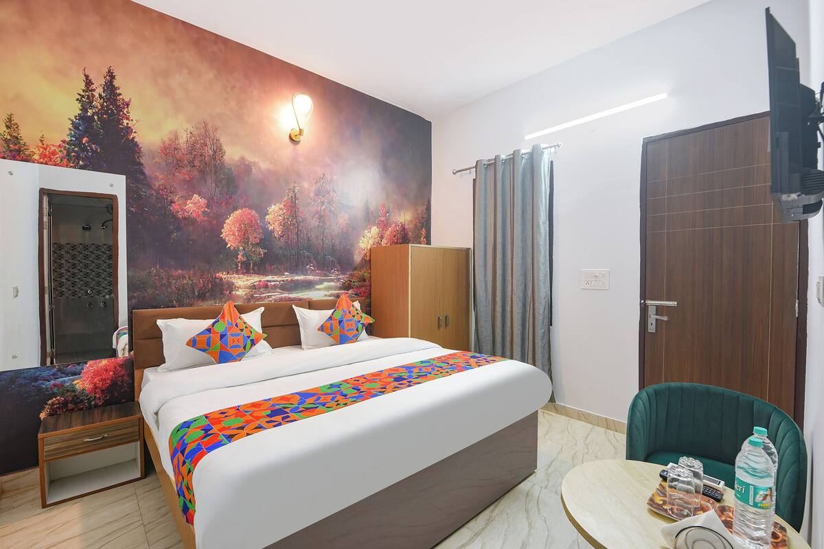 Ein Hotelzimmer mit Bett, Nachttisch, Kleiderschrank, Dusche, einem grünen Sessel, einem Tisch mit Wasserflasche und Fernbedienung sowie einem Wandbild von Waldszenerie.