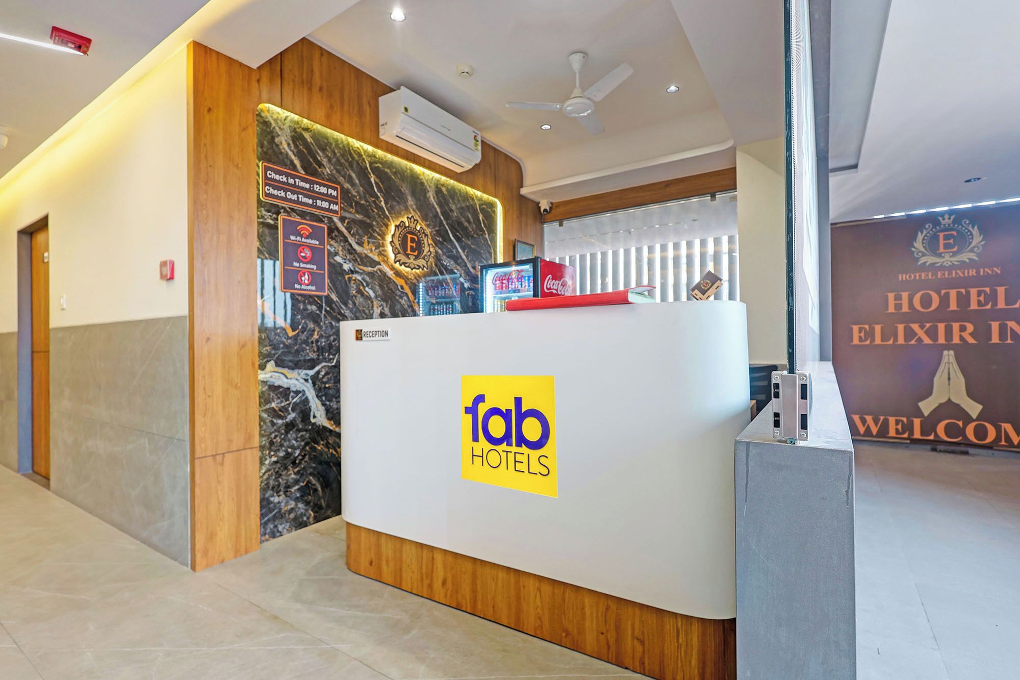 A reception desk with a marble wall and wooden accents, a sign indicating check-in and check-out times, and a welcome sign for Hotel Elixir Inn.