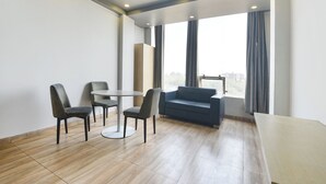 Quarto de hotel moderno com uma mesa redonda, duas poltronas, um sofá e janelas amplas com cortinas.