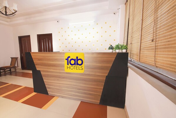 A reception desk with a 'Fab Hotels' sign, a wooden floor with a geometric pattern, a potted plant, and a traditional woven blind on the window. - Fabhotel 9 Palm (Dehradun)