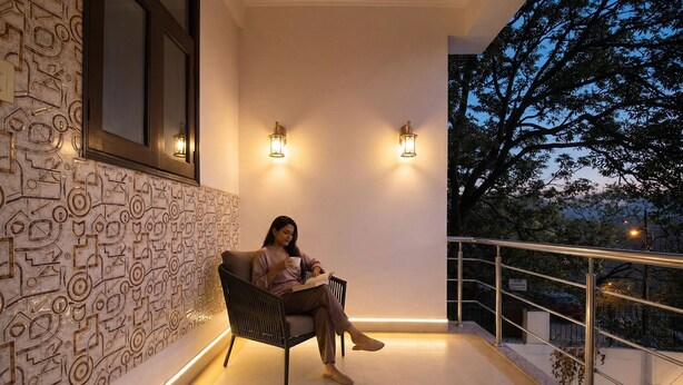 A person sitting on a bench on a balcony with patterned wall and two wall-mounted lights. - Fabhotel 9 Palm (Dehradun)