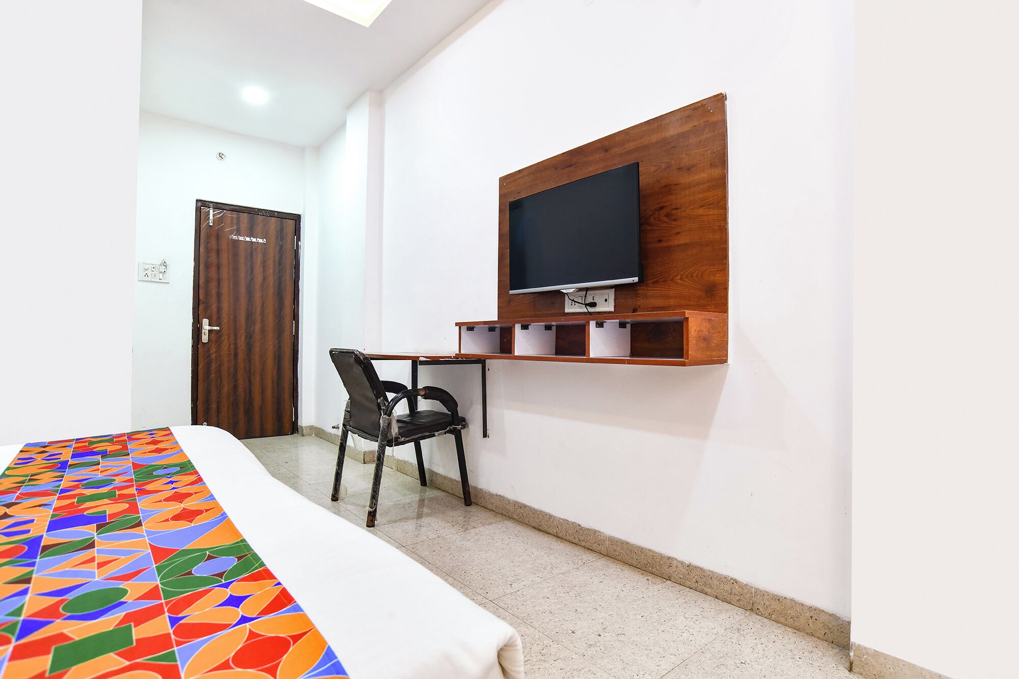 A hotel room with a bed, a desk with a chair, a television mounted on the wall, and a door.