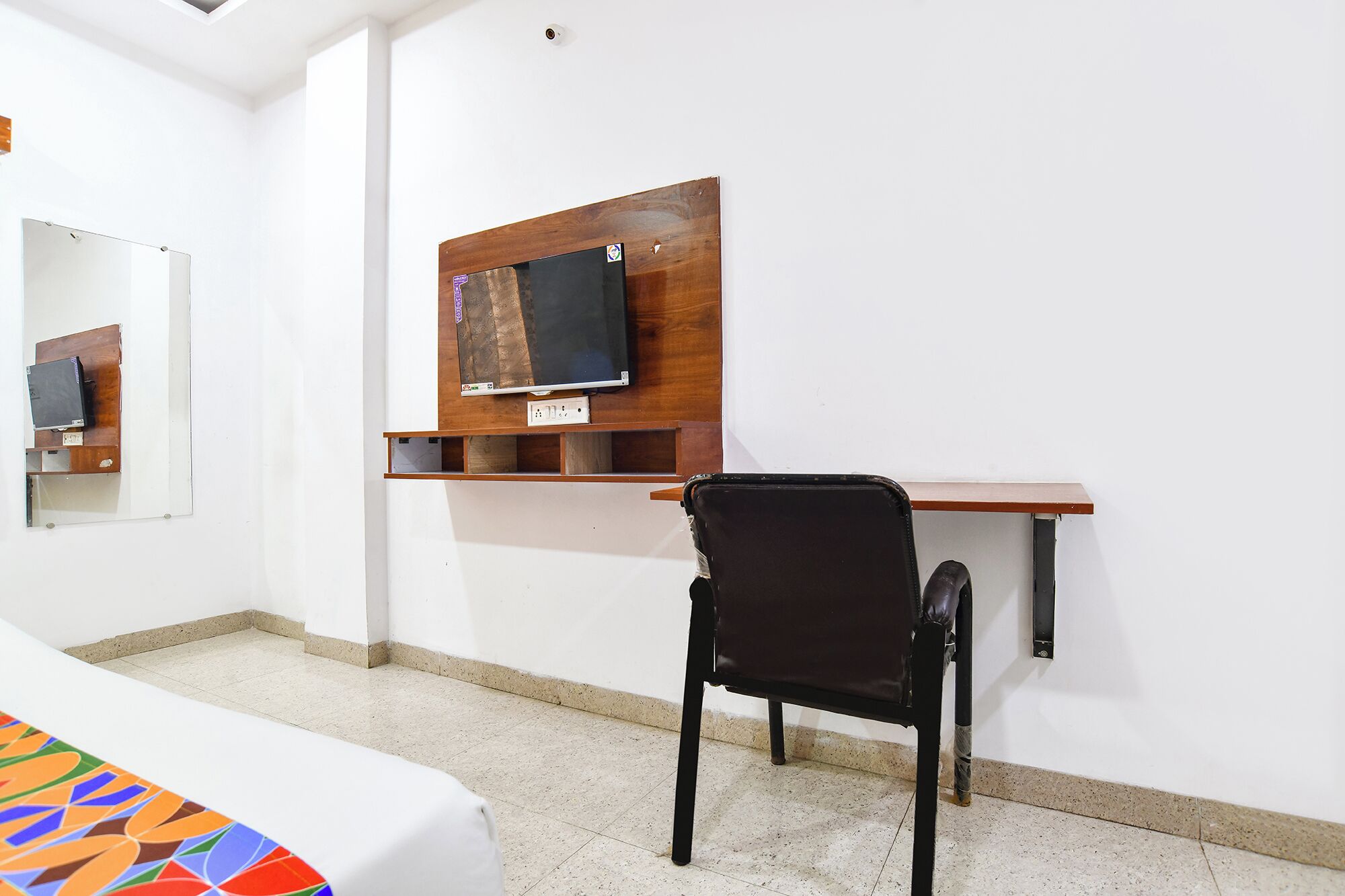 A room with a bed, a desk with a chair, a television mounted on a wooden shelf, and a mirror on the wall.