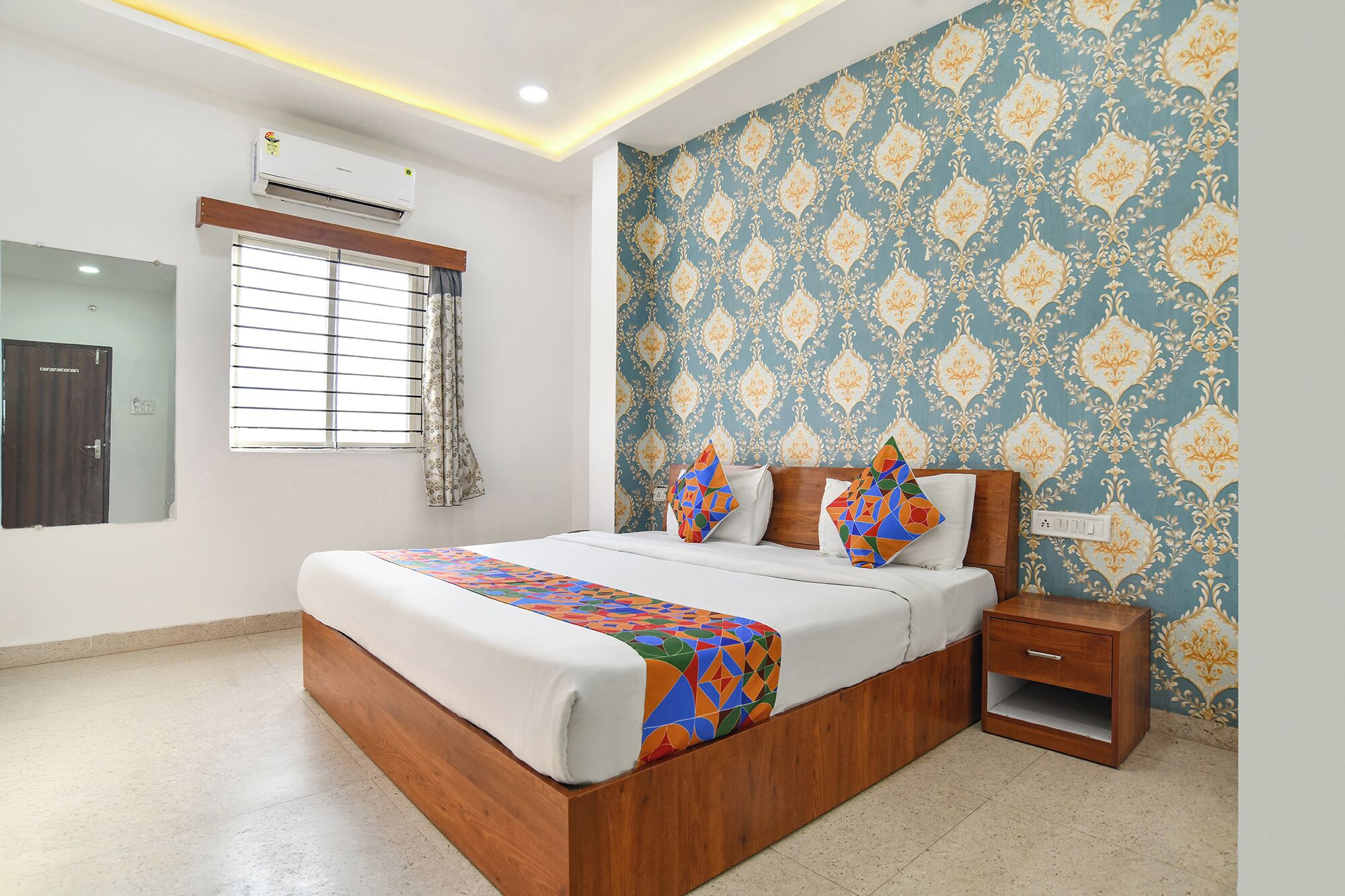 A bedroom with a wooden bed, a nightstand, a wall-mounted air conditioner, a window with blinds, and a patterned wallpaper.