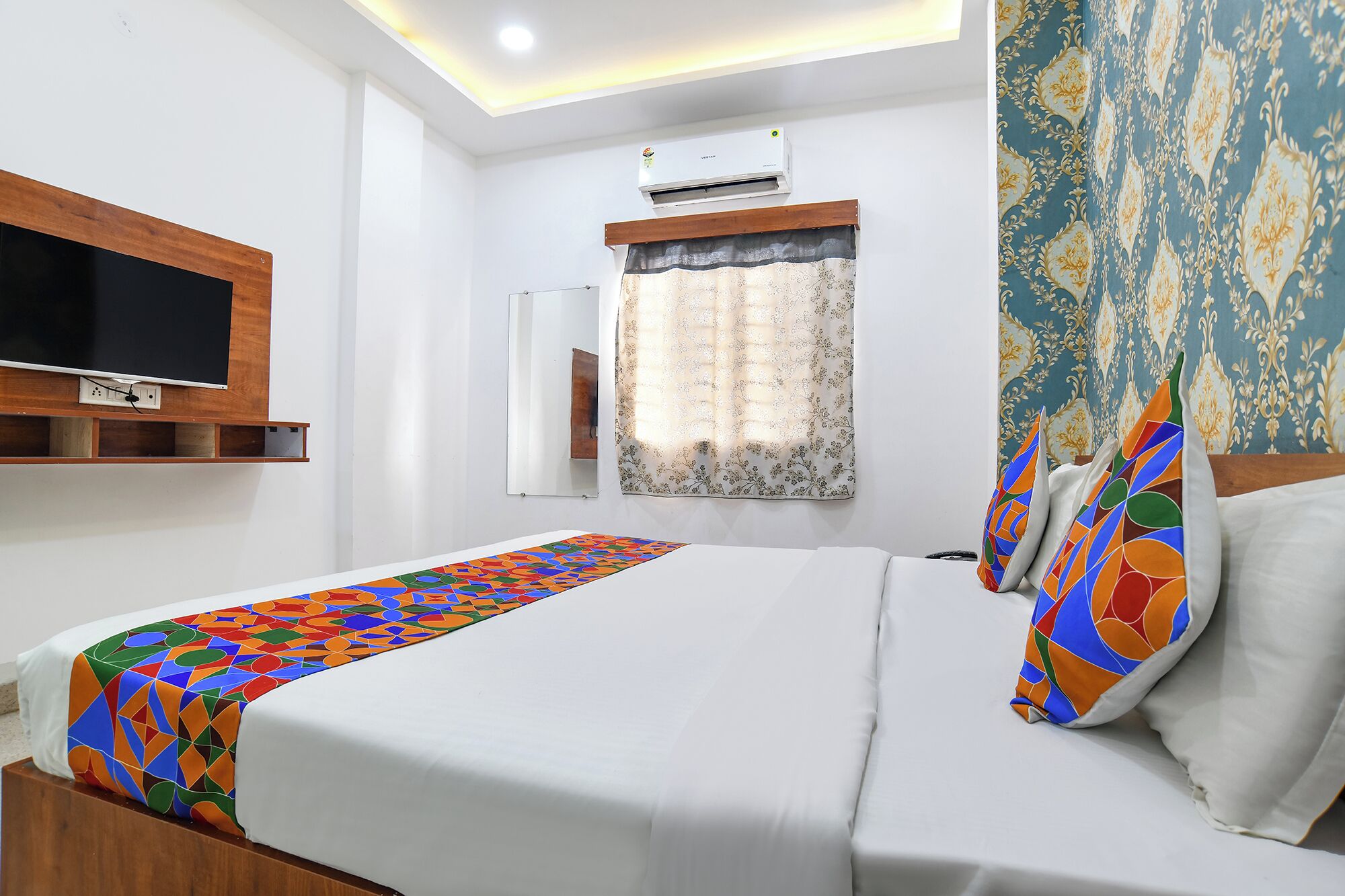 A hotel room with a bed, a TV mounted on the wall, an air conditioning unit, a window with curtains, and a patterned wall.