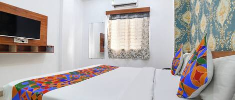 A hotel room with a bed, a TV mounted on the wall, an air conditioning unit, a window with curtains, and a patterned wall.