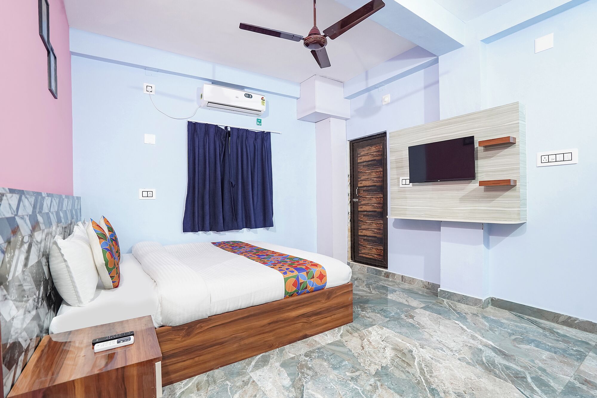 A bedroom with a bed, a ceiling fan, an air conditioner, a TV mounted on the wall, a wooden door, and a marble floor.