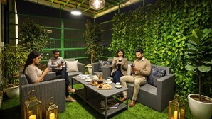 A group of people seated on wicker sofas around a coffee table with food and drinks, in a room with a green leafy wall and artificial grass flooring. - Fabhotel Starex Inn (Gurugram)
