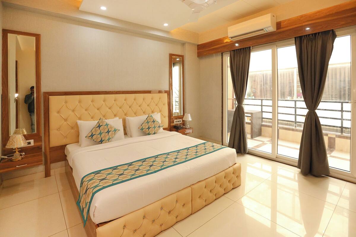 Une chambre d’hôtel avec un grand lit, un miroir, une table de chevet avec une lampe et une vue sur l’extérieur grâce à de grandes fenêtres.
