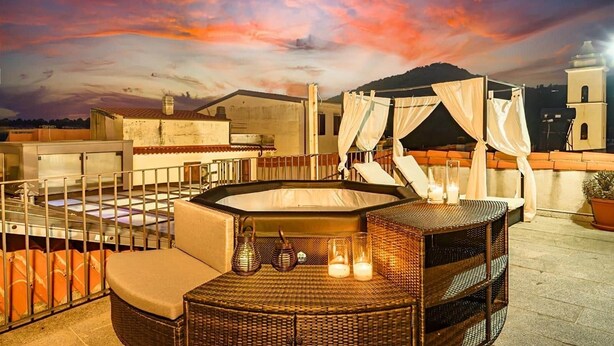 A rooftop terrace with a hot tub, wicker furniture, and a view of the cityscape at sunset.