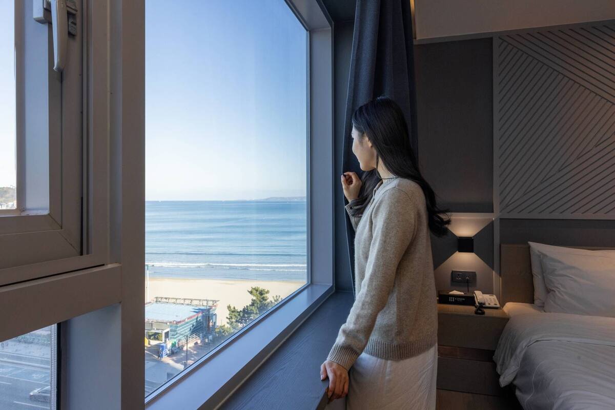 Une chambre d’hôtel dotée d’une grande fenêtre donnant sur une plage et l’océan, d’un lit, d’une table de chevet avec une lampe et d’un téléphone.