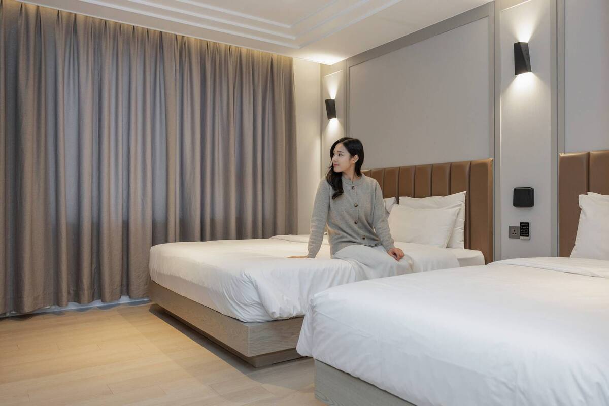 Une chambre d’hôtel avec deux lits, une grande fenêtre avec des rideaux et un luminaire moderne.