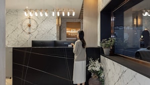 Un comptoir d’accueil d’hôtel avec un mur en marbre, un lustre moderne et une plante en pot.
