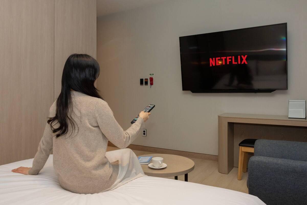 Une personne est assise sur un lit dans une chambre d’hôtel, tenant une télécommande, et un téléviseur affiche le mot « NETFLIX » juste au-dessus.