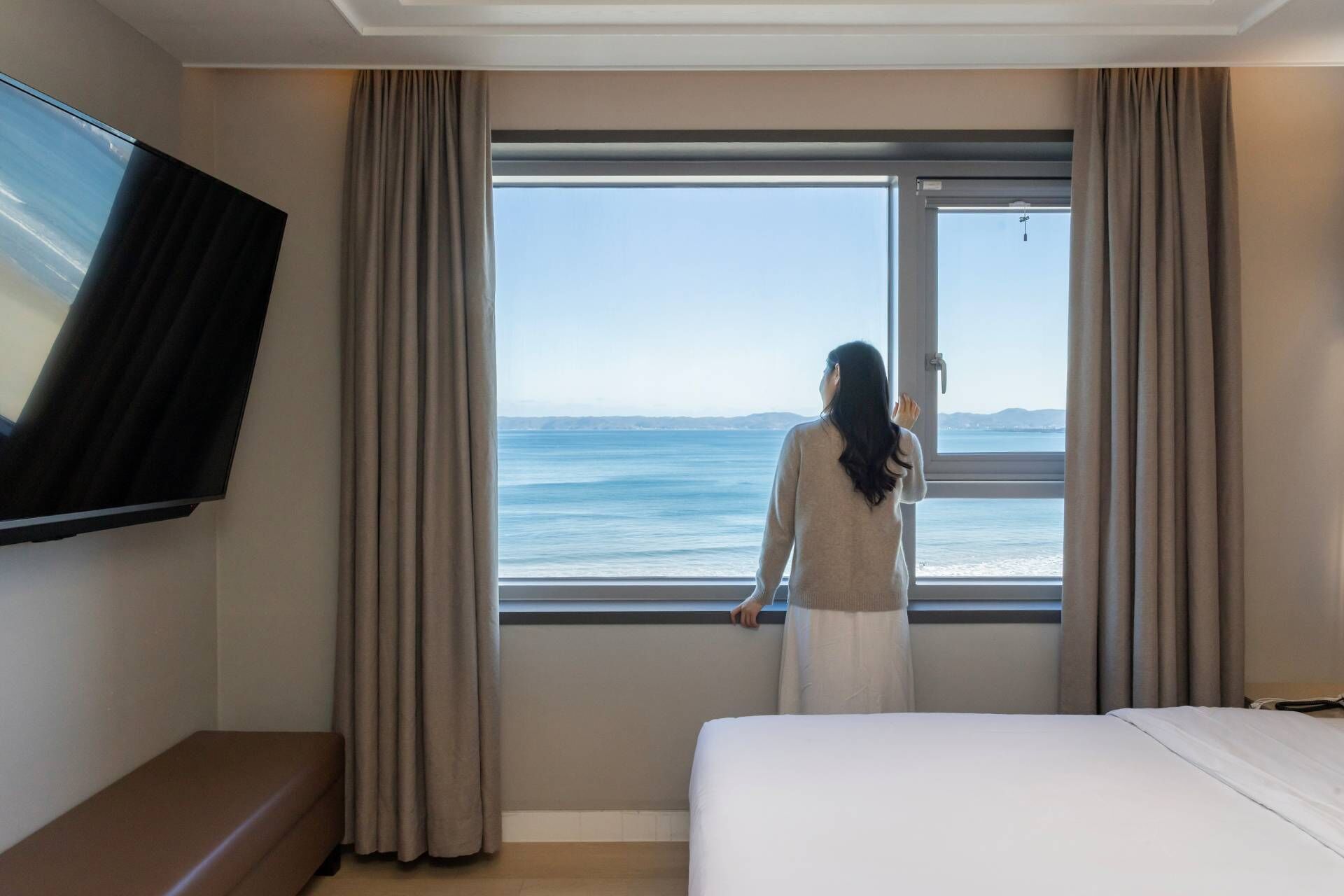 Habitación de hotel con una ventana grande que ofrece vistas al mar, una cama y un televisor de pantalla plana montado en la pared.