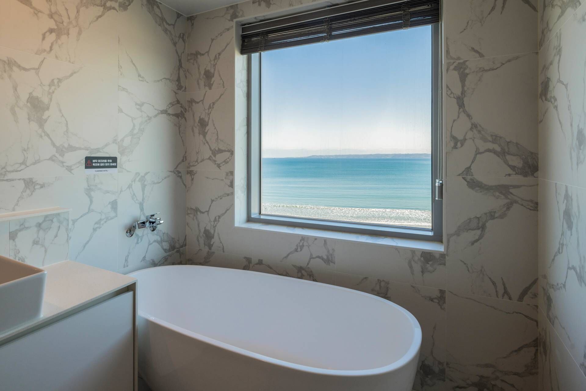 A bathroom with a bathtub, a window with a view of the sea, and marble-patterned walls.
