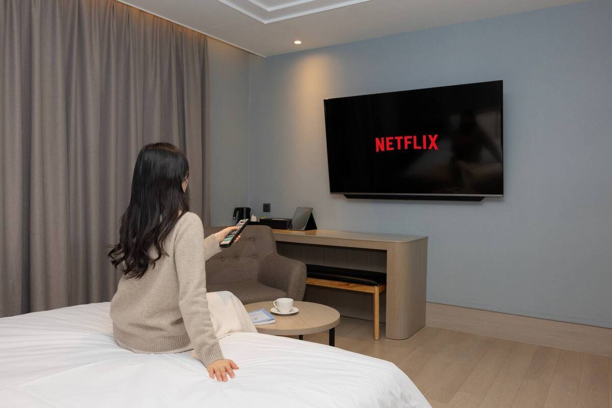 Une personne est assise sur un lit dans une chambre d’hôtel, face à un téléviseur qui affiche le logo de Netflix.