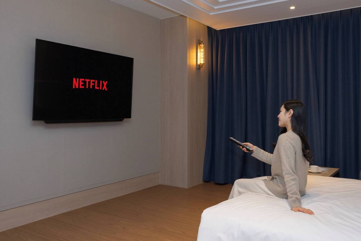 Une chambre d’hôtel dotée d’un grand téléviseur à écran plat affichant le logo de Netflix, d’une lampe fixée au mur et d’un lit recouvert d’une couette blanche.