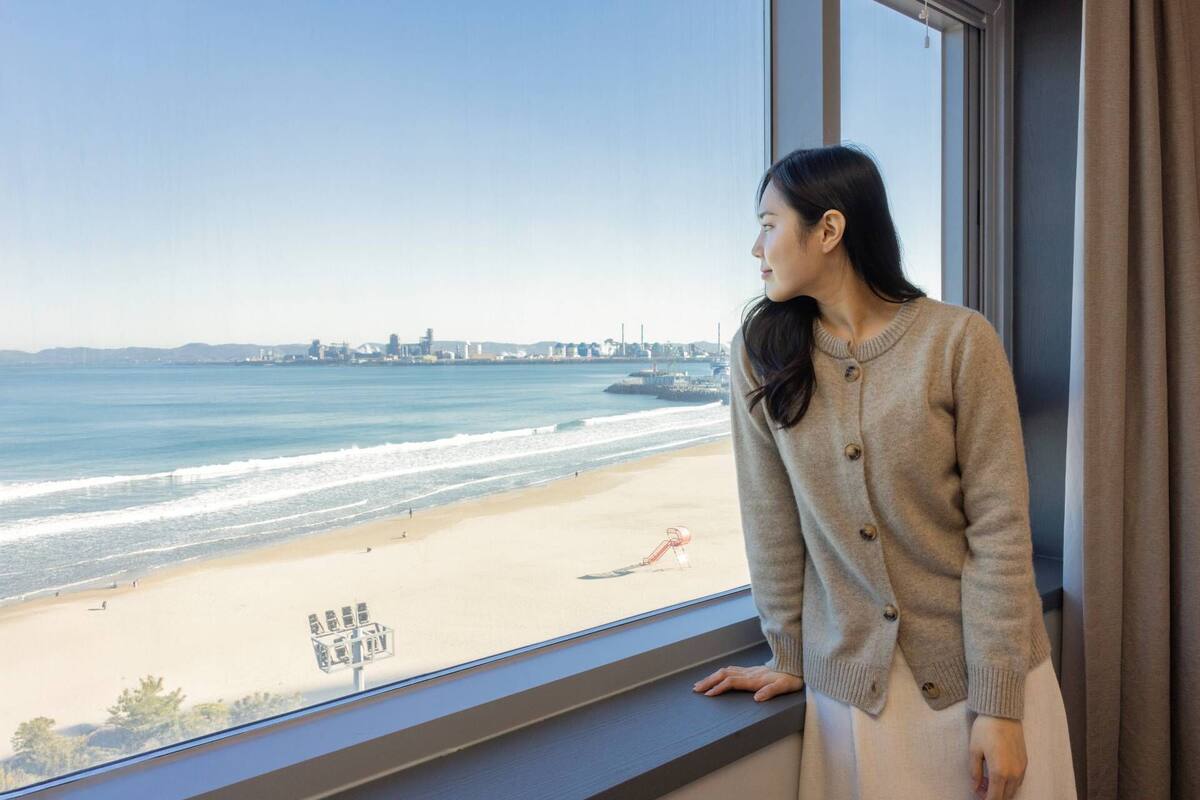 Une personne appuyée à une fenêtre donnant sur une plage, avec une vue sur les bâtiments d’une ville au loin.
