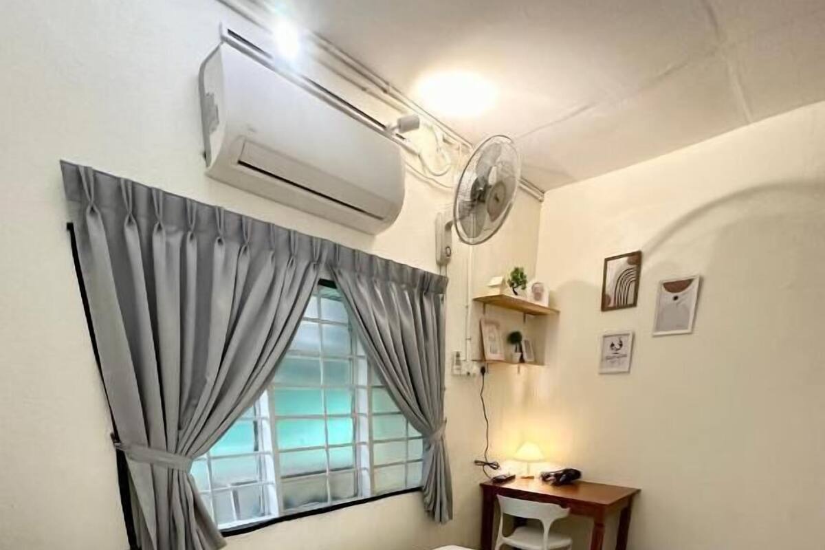 A hotel room with a bed, pillows, a desk with a lamp, a fan, and an air conditioning unit.