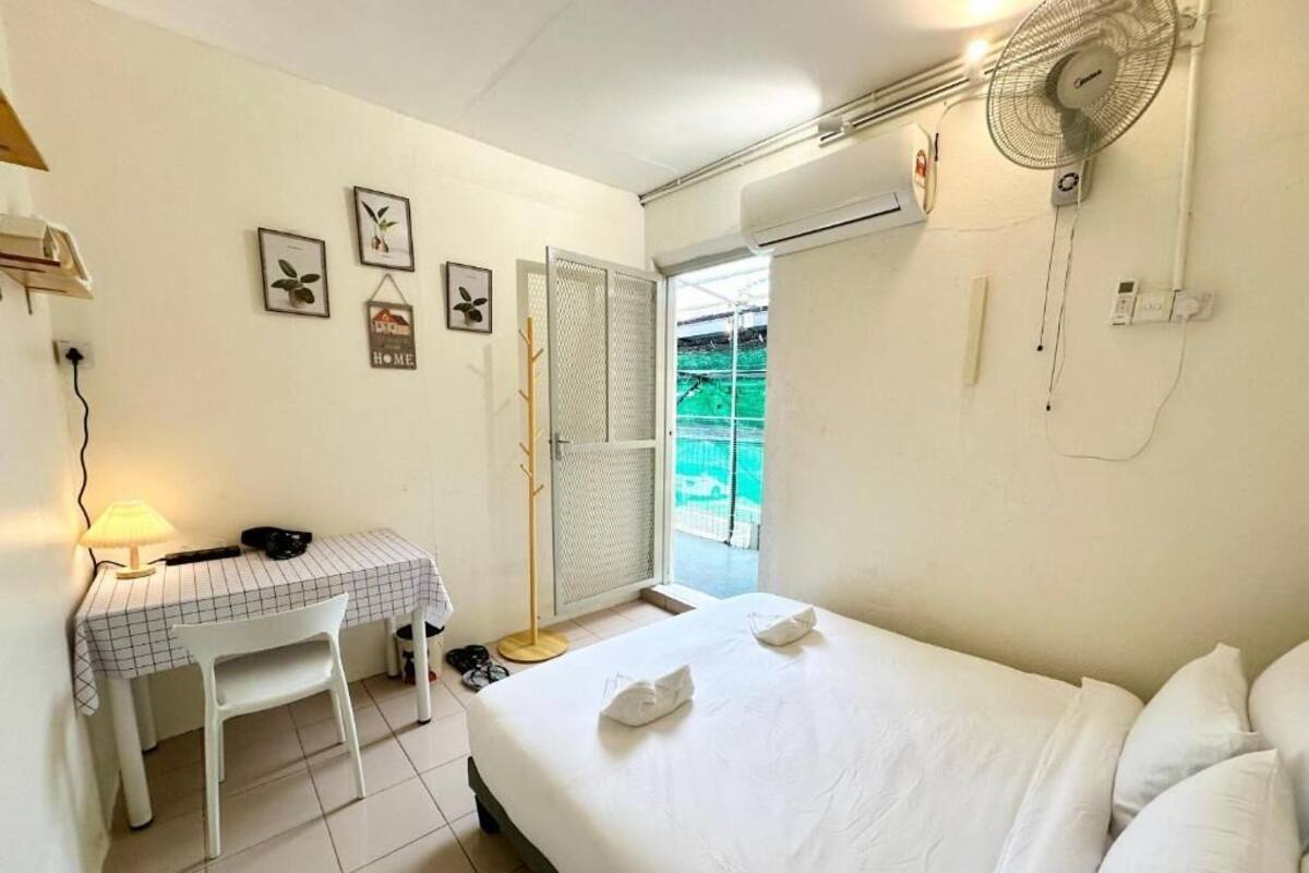 A bedroom with a bed, a desk with a chair, a standing fan, and a window with a view of greenery.
