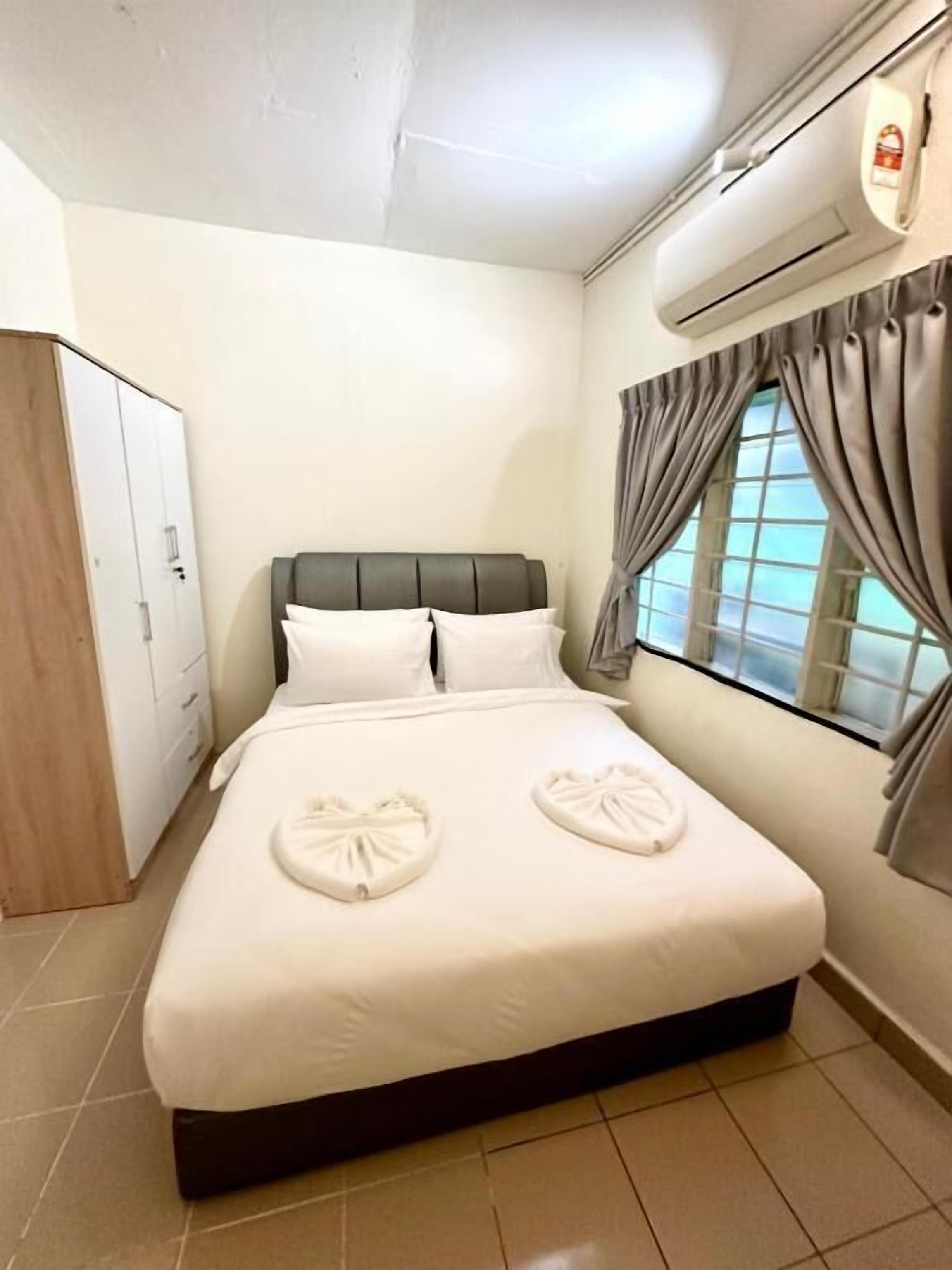 A neatly made bed with white linens and two heart-shaped towels, a wardrobe, and a window with curtains.