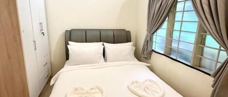 A neatly made bed with white linens and two heart-shaped towels, a wardrobe, and a window with curtains.