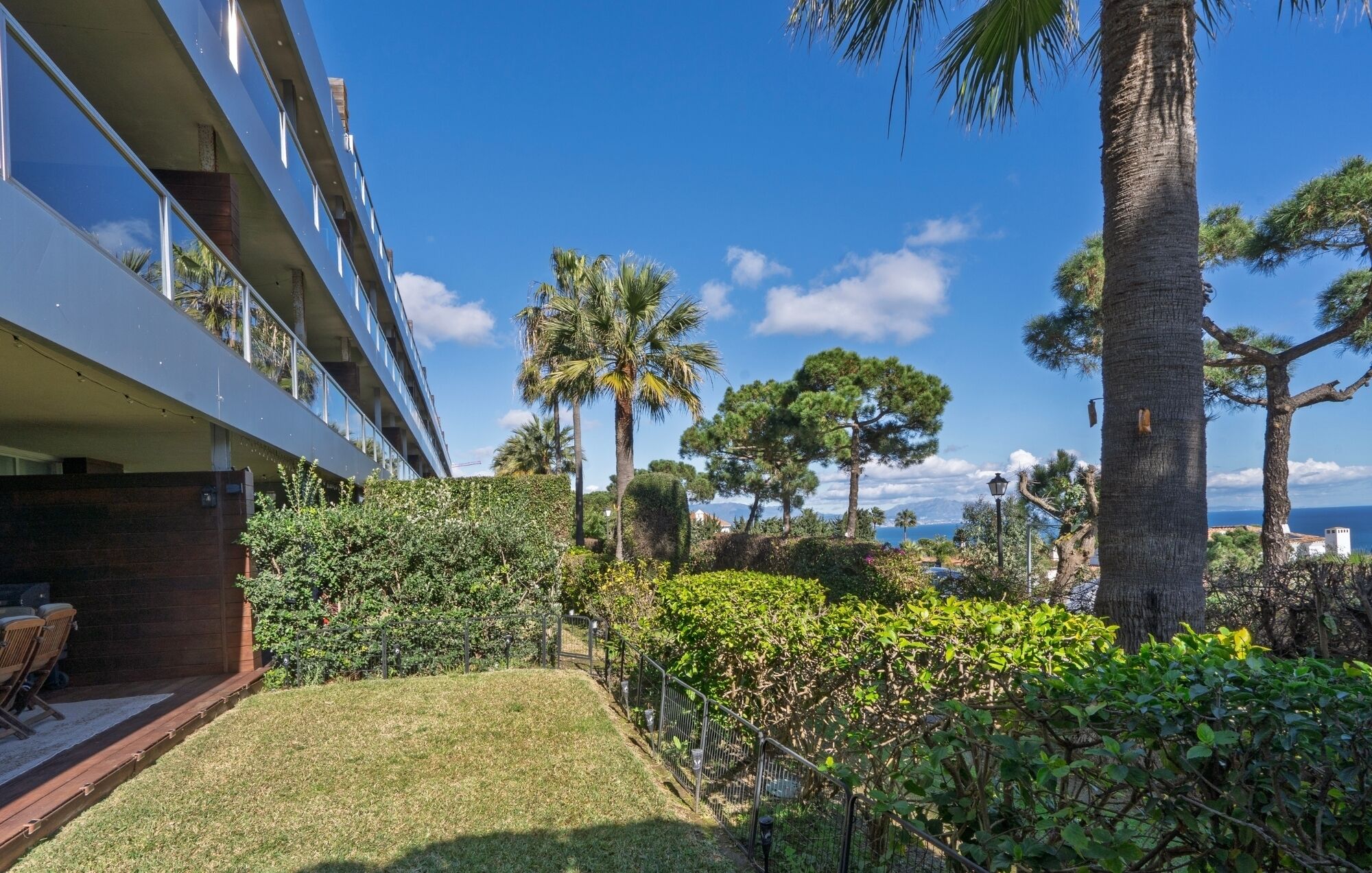 A modern building with a balcony overlooking a landscaped garden with palm trees and a view of the sea.