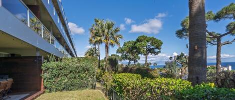 A modern building with a balcony overlooking a landscaped garden with palm trees and a view of the sea.