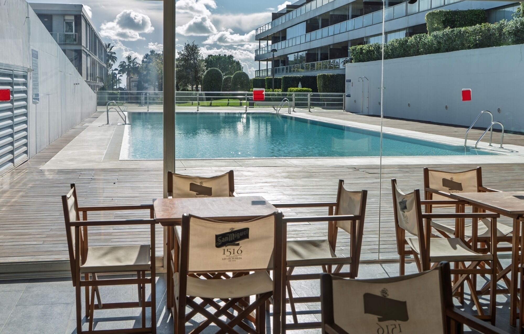 A poolside area with wooden chairs and tables, a clear view of the pool, and a modern building in the background.
