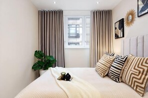 A hotel room with a bed, curtains, a window, a plant, and a wall decoration. - One Bed Serviced Apt Near Holborn in Chancery Lane (London)