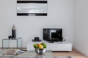 Un salon moderne avec un téléviseur à écran plat, un meuble de rangement blanc, une table basse en verre sur laquelle se trouve un saladier rempli de fruits, et un magazine.