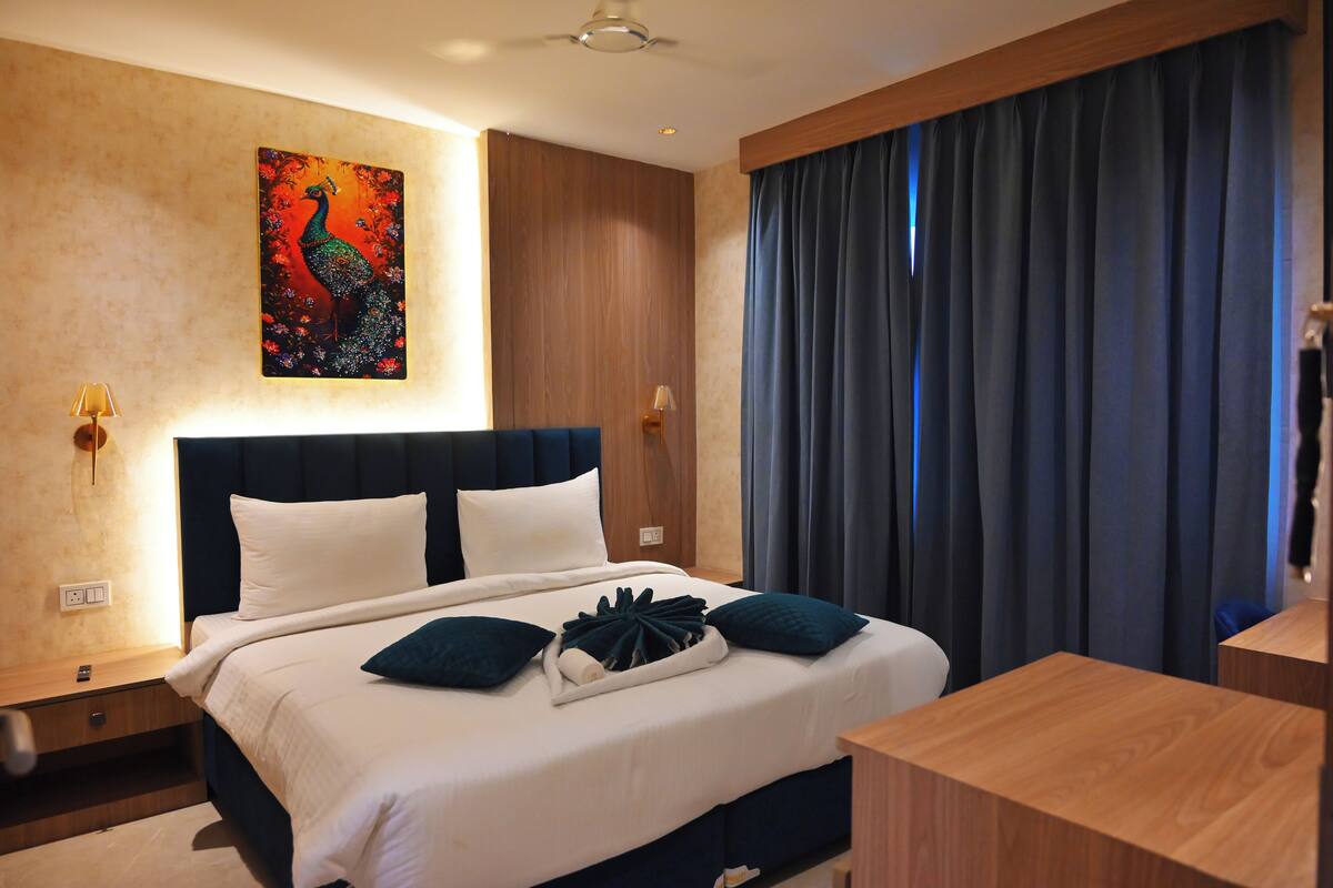 Une chambre d’hôtel avec un grand lit, un tableau représentant un paon accroché au mur et une vue sur le ciel nocturne, visible à travers d’épais rideaux.