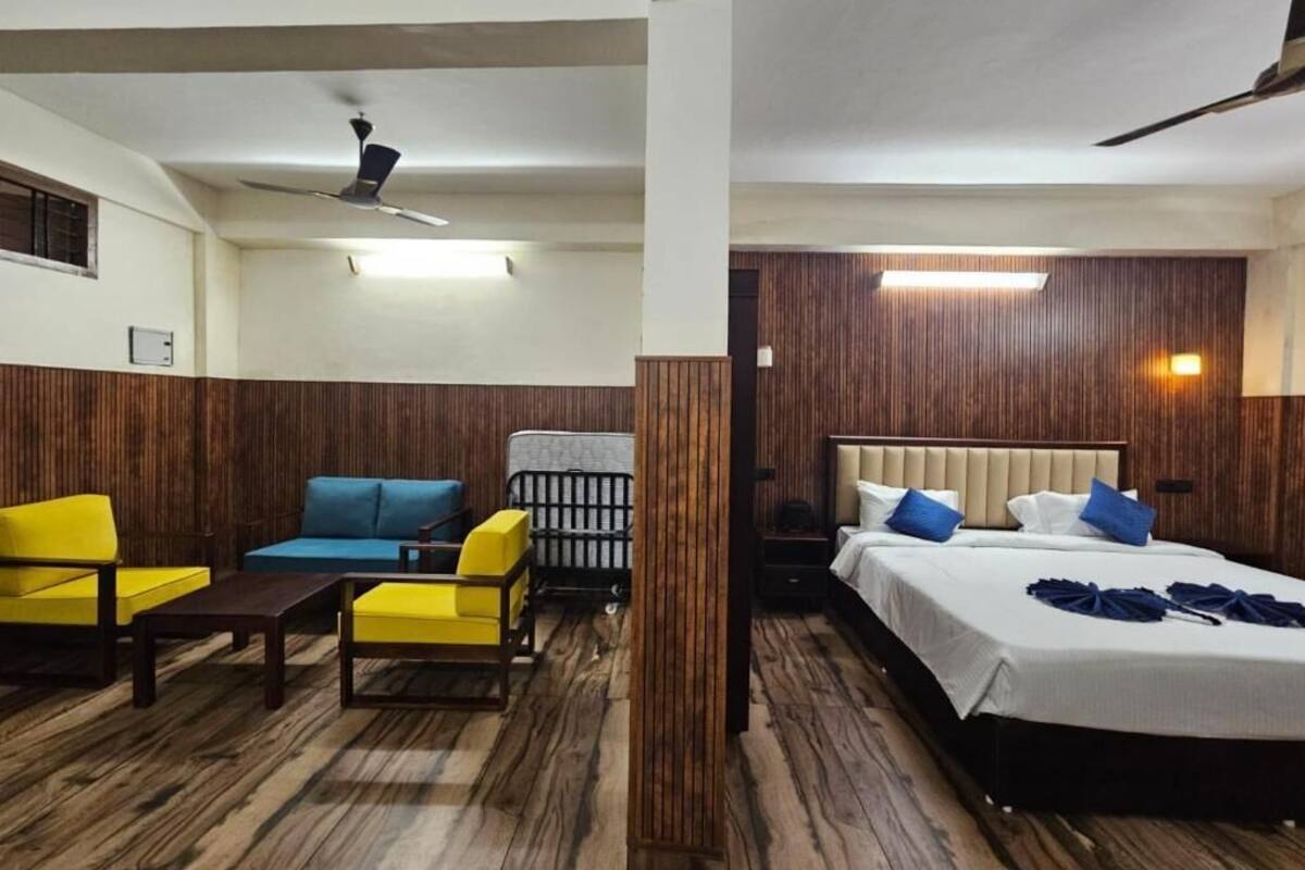 A hotel room with a bed, a sofa, a chair, and a small table. There is a ceiling fan and a wall-mounted light.