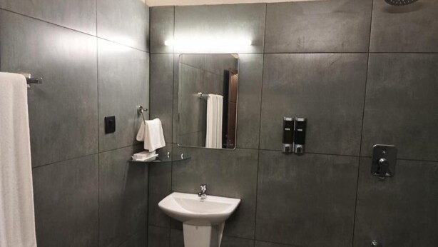 A modern bathroom with dark tiles, a white sink, a mirror, and a shower area. - The Vintage Haven Munnar (Udumbanchola)