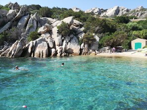 A beach with clear turquoise water, rocky cliffs, and a small building with a green door.